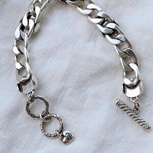 May include: A silver chain bracelet with large, interlocking links. The bracelet features a toggle clasp and decorative rings with a heart-shaped charm. The jewelry is displayed on a white, textured surface.