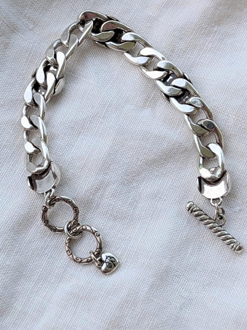 May include: A silver chain bracelet with large, interlocking links. The bracelet features a toggle clasp and decorative rings with a heart-shaped charm. The jewelry is displayed on a white, textured surface.