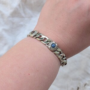 May include: Silver-toned chain bracelet with decorative circular links. Each link features a central blue stone and surrounding floral design. The bracelet is worn on a wrist, showcasing its chunky, textured design.