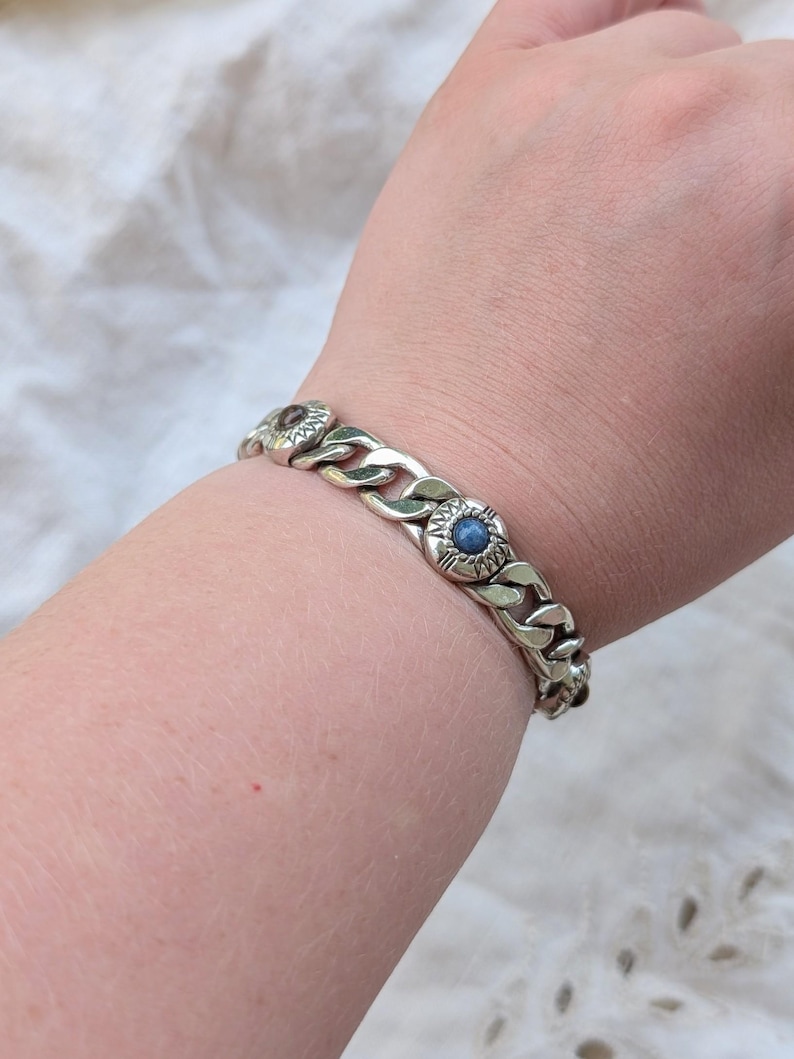May include: Silver-toned chain bracelet with decorative circular links. Each link features a central blue stone and surrounding floral design. The bracelet is worn on a wrist, showcasing its chunky, textured design.