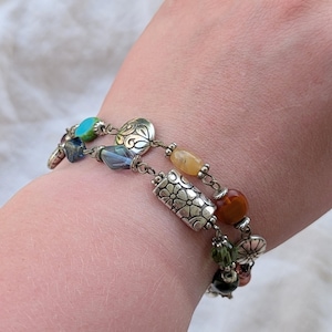 K&ouml;nnte beinhalten: Ein mehrstr&auml;ngiges Armband mit silberfarbenen Metallperlen mit Blumen- und Wirbelmustern, durchsetzt mit verschiedenen bunten Glas- und Steinperlen in T&uuml;rkis, Blau, Gelb, Orange, Gr&uuml;n und Dunkelbraun an einer Metallkette.