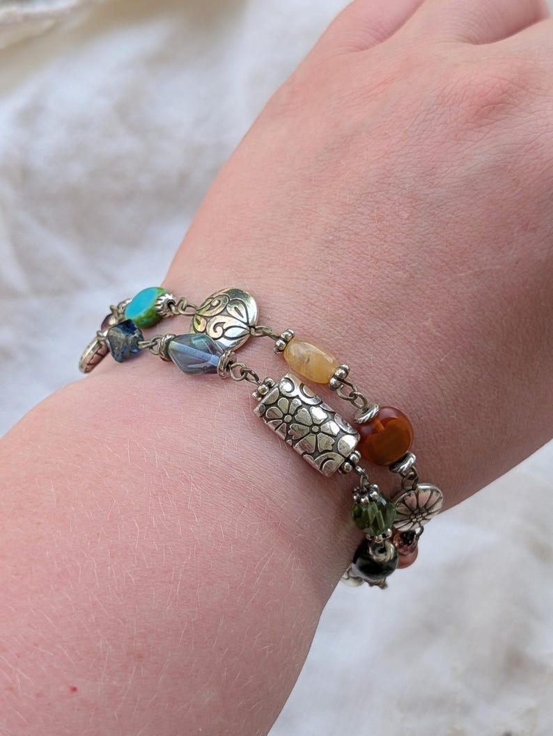K&ouml;nnte beinhalten: Ein mehrstr&auml;ngiges Armband mit silberfarbenen Metallperlen mit Blumen- und Wirbelmustern, durchsetzt mit verschiedenen bunten Glas- und Steinperlen in T&uuml;rkis, Blau, Gelb, Orange, Gr&uuml;n und Dunkelbraun an einer Metallkette.