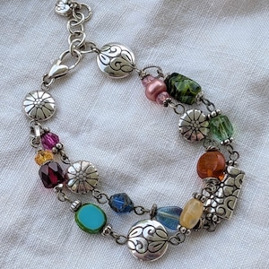 K&ouml;nnte beinhalten: Ein mehrstr&auml;ngiges silberfarbenes Bettelarmband mit einer Vielzahl bunter Glas- und Kristallperlen in Rosa, Gr&uuml;n, Blau, Gelb und Bernstein, durchsetzt mit runden Metallanh&auml;ngern, die mit Blumen- und Schn&ouml;rkelmustern verziert sind.