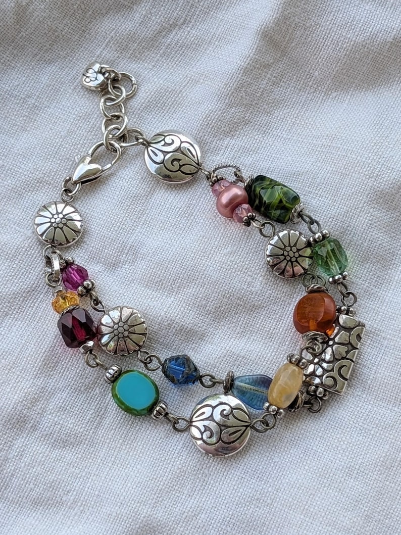 K&ouml;nnte beinhalten: Ein mehrstr&auml;ngiges silberfarbenes Bettelarmband mit einer Vielzahl bunter Glas- und Kristallperlen in Rosa, Gr&uuml;n, Blau, Gelb und Bernstein, durchsetzt mit runden Metallanh&auml;ngern, die mit Blumen- und Schn&ouml;rkelmustern verziert sind.