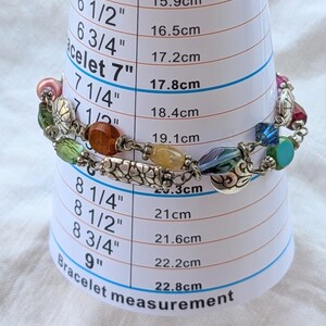 K&ouml;nnte beinhalten: Ein mehrfarbiges Perlenarmband mit silberfarbenen Metallanh&auml;ngern und verschiedenen Glasperlen in Rosa, Gr&uuml;n, Orange, Gelb und Blau, platziert auf einem wei&szlig;en Messkegel, der eine Gr&ouml;&szlig;e von etwa 19,1 cm anzeigt.