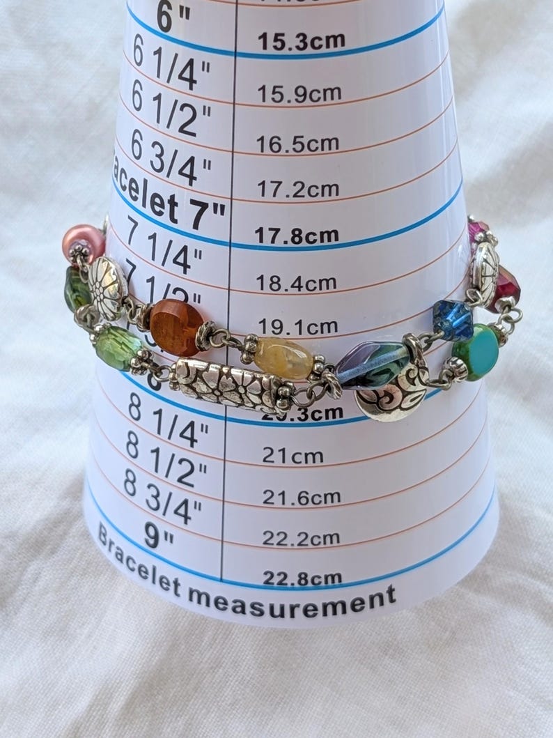 K&ouml;nnte beinhalten: Ein mehrfarbiges Perlenarmband mit silberfarbenen Metallanh&auml;ngern und verschiedenen Glasperlen in Rosa, Gr&uuml;n, Orange, Gelb und Blau, platziert auf einem wei&szlig;en Messkegel, der eine Gr&ouml;&szlig;e von etwa 19,1 cm anzeigt.