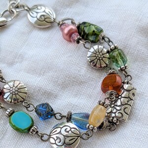 K&ouml;nnte beinhalten: Ein mehrstr&auml;ngiges silberfarbenes Armband mit einer Vielzahl bunter Glasperlen und Metallanh&auml;ngern. Die Perlen in Rosa, Gr&uuml;n, Orange, Gelb und Blau sind mit runden silbernen Scheiben und einem rechteckigen Blumenanh&auml;nger kombiniert.