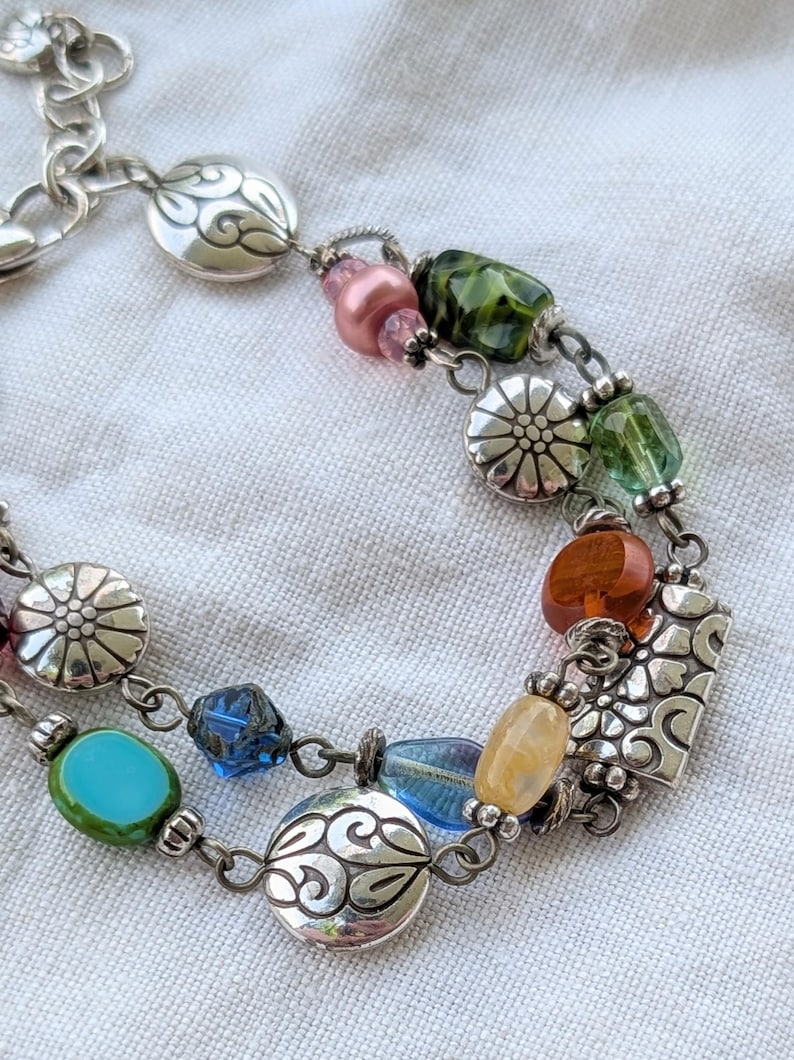 K&ouml;nnte beinhalten: Ein mehrstr&auml;ngiges silberfarbenes Armband mit einer Vielzahl bunter Glasperlen und Metallanh&auml;ngern. Die Perlen in Rosa, Gr&uuml;n, Orange, Gelb und Blau sind mit runden silbernen Scheiben und einem rechteckigen Blumenanh&auml;nger kombiniert.