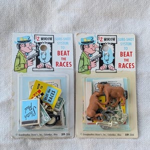 May include: Two vintage horse racing game sets. Each set includes a plastic blister pack with a cartoon illustration of a man with a gun and the text "$2 Window Sure-Shot System to Beat the Races". One set includes a sketch pad and play money.
