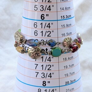 K&ouml;nnte beinhalten: Ein mehrstr&auml;ngiges Armband mit silberfarbenen Metallanh&auml;ngern mit komplexen Mustern, durchsetzt mit facettierten blauen, gr&uuml;nen, gelben und violetten Glasperlen. Das Armband ist auf einem Messkegel mit einem Umfang von 17,1 cm dargestellt.