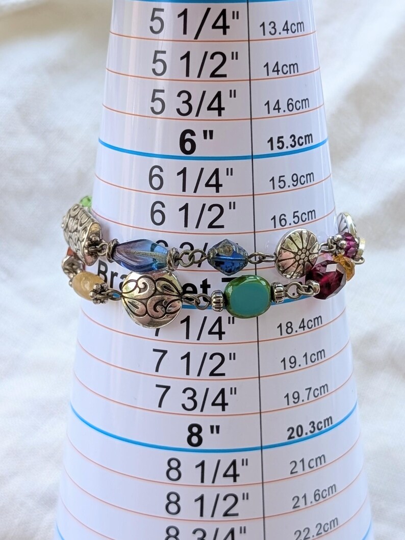 K&ouml;nnte beinhalten: Ein mehrstr&auml;ngiges Armband mit silberfarbenen Metallanh&auml;ngern mit komplexen Mustern, durchsetzt mit facettierten blauen, gr&uuml;nen, gelben und violetten Glasperlen. Das Armband ist auf einem Messkegel mit einem Umfang von 17,1 cm dargestellt.