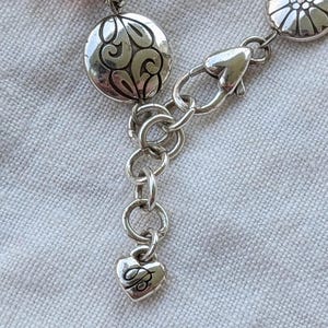 K&ouml;nnte beinhalten: Ein silberfarbenes Armband mit Karabinerverschluss, runden Metallanh&auml;ngern mit gepr&auml;gten Blumen- und Schn&ouml;rkelmustern sowie einem kleinen herzf&ouml;rmigen Anh&auml;nger am Ende der Verl&auml;ngerungskette, in den der Buchstabe B eingraviert ist.