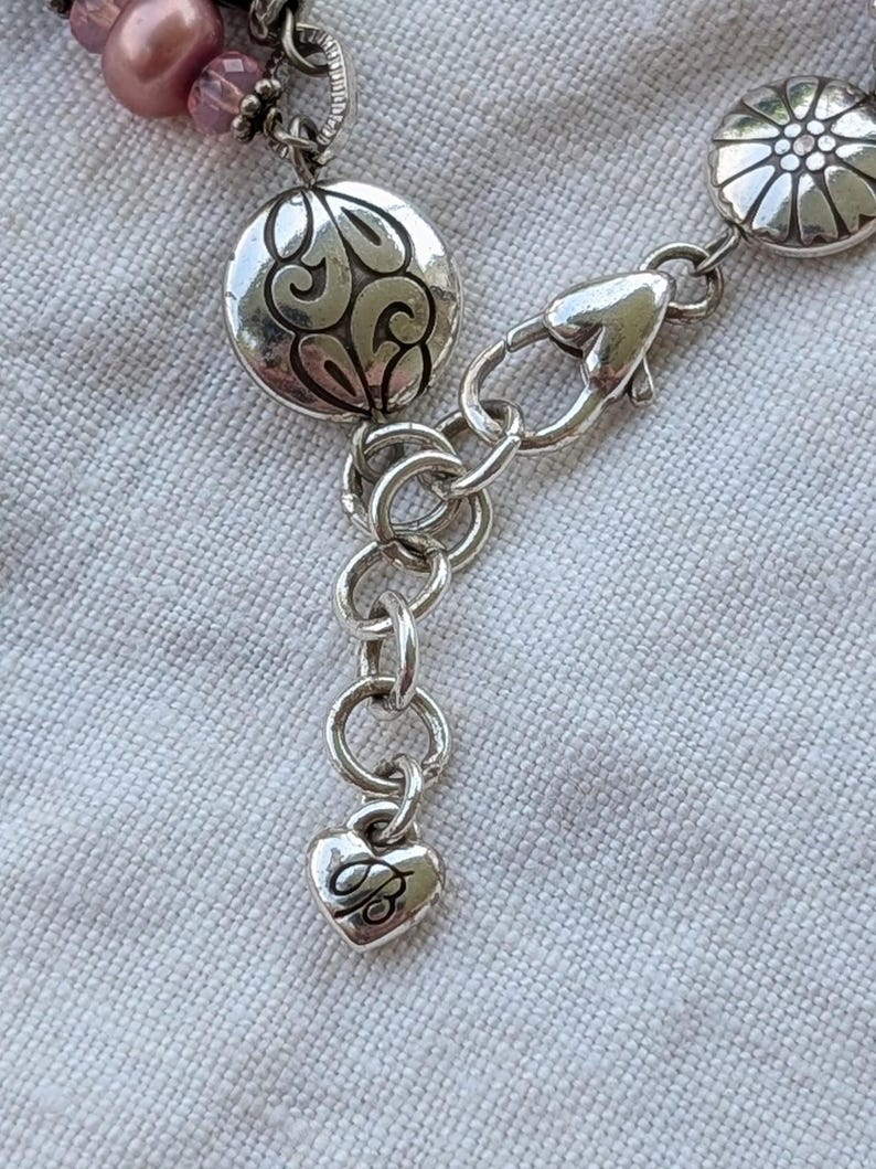 K&ouml;nnte beinhalten: Ein silberfarbenes Armband mit Karabinerverschluss, runden Metallanh&auml;ngern mit gepr&auml;gten Blumen- und Schn&ouml;rkelmustern sowie einem kleinen herzf&ouml;rmigen Anh&auml;nger am Ende der Verl&auml;ngerungskette, in den der Buchstabe B eingraviert ist.