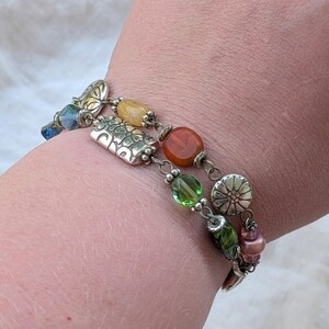 K&ouml;nnte beinhalten: Ein mehrreihiges Armband mit einer Vielzahl bunter Perlen und silberfarbenen Metallanh&auml;ngern. Das Design umfasst durchscheinende gr&uuml;ne, orangefarbene und blaue Perlen sowie strukturierte silberfarbene Metallzwischenst&uuml;cke und bl&uuml;tenf&ouml;rmige Akzente.