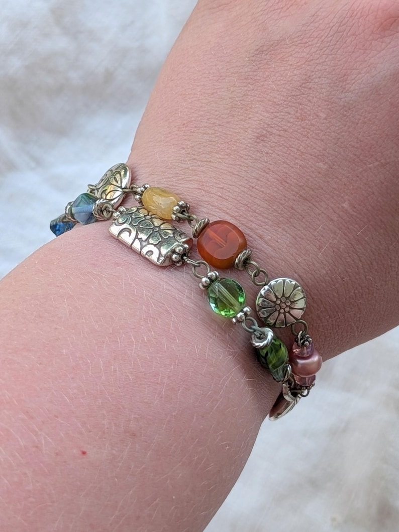 K&ouml;nnte beinhalten: Ein mehrreihiges Armband mit einer Vielzahl bunter Perlen und silberfarbenen Metallanh&auml;ngern. Das Design umfasst durchscheinende gr&uuml;ne, orangefarbene und blaue Perlen sowie strukturierte silberfarbene Metallzwischenst&uuml;cke und bl&uuml;tenf&ouml;rmige Akzente.