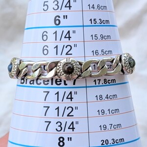 May include: A silver-toned bracelet with a chain link design, featuring decorative circular elements with dark accents. The bracelet is measured on a sizing cone, appearing to be approximately 7 inches in length.