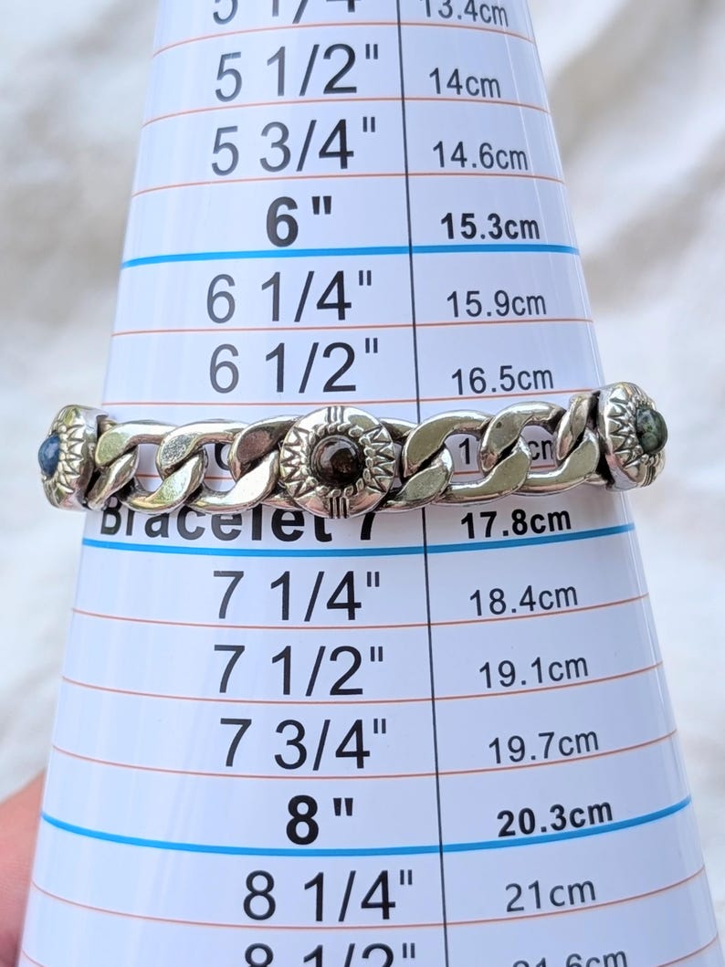 May include: A silver-toned bracelet with a chain link design, featuring decorative circular elements with dark accents. The bracelet is measured on a sizing cone, appearing to be approximately 7 inches in length.