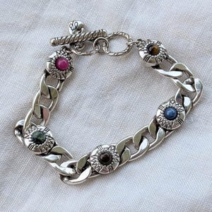 May include: A silver chain bracelet with a toggle clasp. The bracelet features five decorative circular settings, each with a different colored gemstone: pink, brown, blue, green, and tiger's eye. The bracelet is displayed on a white fabric surface.