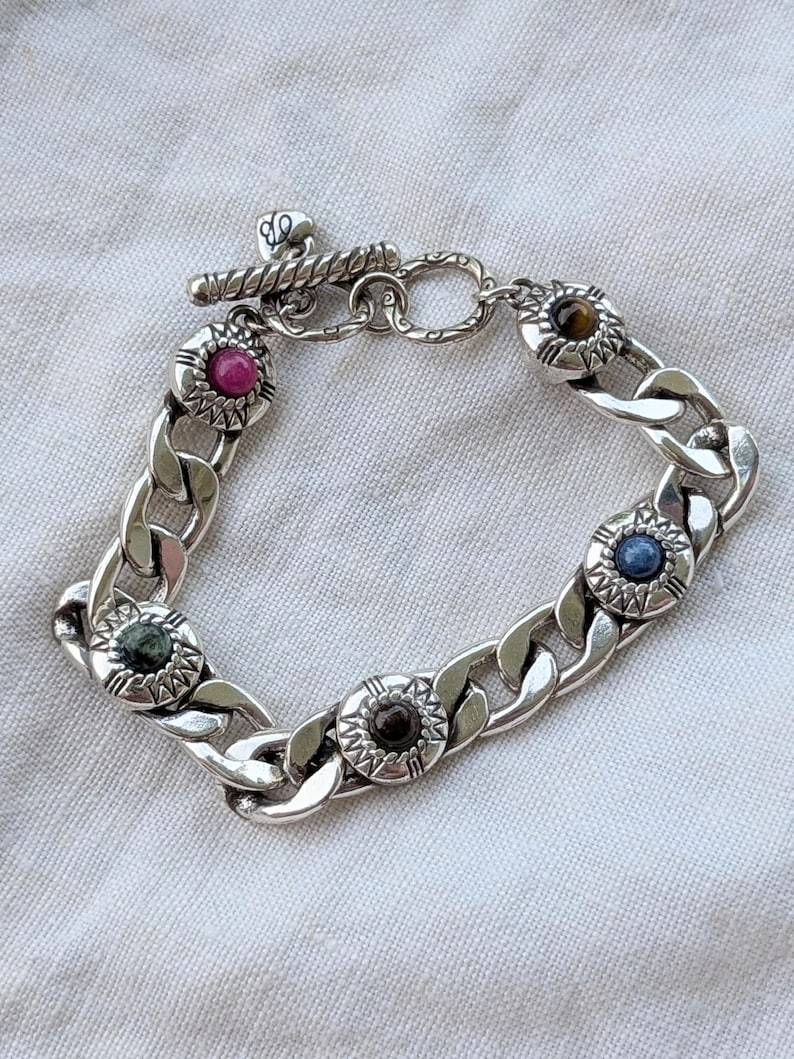 May include: A silver chain bracelet with a toggle clasp. The bracelet features five decorative circular settings, each with a different colored gemstone: pink, brown, blue, green, and tiger's eye. The bracelet is displayed on a white fabric surface.