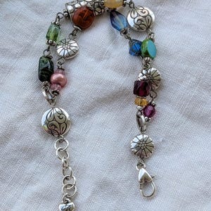 K&ouml;nnte beinhalten: Ein mehrstr&auml;ngiges Bettelarmband mit silberfarbenen Metallgliedern, blumenmuster-Scheiben und einer Auswahl an bunten Glas- und Kristallperlen in Blau, Gr&uuml;n, Rot, Rosa und Gelb, abgerundet mit einem herzf&ouml;rmigen Karabinerverschluss.