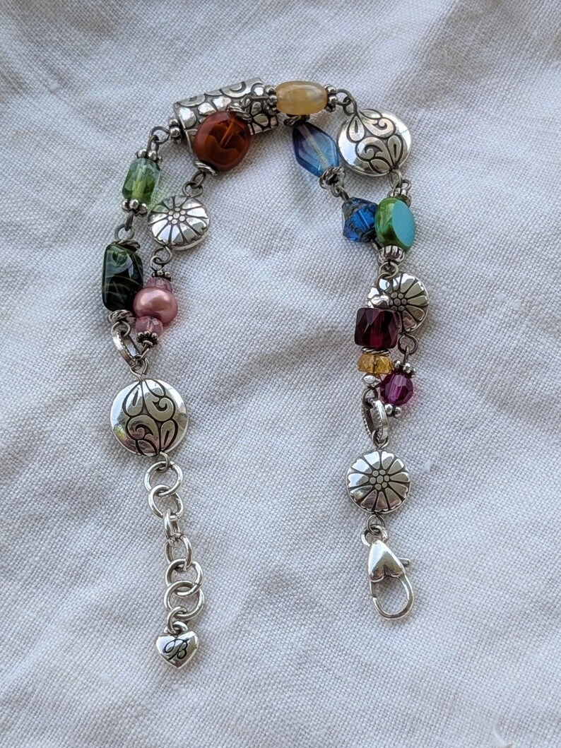 K&ouml;nnte beinhalten: Ein mehrstr&auml;ngiges Bettelarmband mit silberfarbenen Metallgliedern, blumenmuster-Scheiben und einer Auswahl an bunten Glas- und Kristallperlen in Blau, Gr&uuml;n, Rot, Rosa und Gelb, abgerundet mit einem herzf&ouml;rmigen Karabinerverschluss.