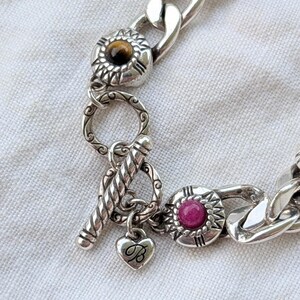May include: Silver-toned bracelet with a chunky chain design. The bracelet features decorative circular accents with a brown and a pink stone, and a heart charm with the letter "B". The clasp is a toggle style.