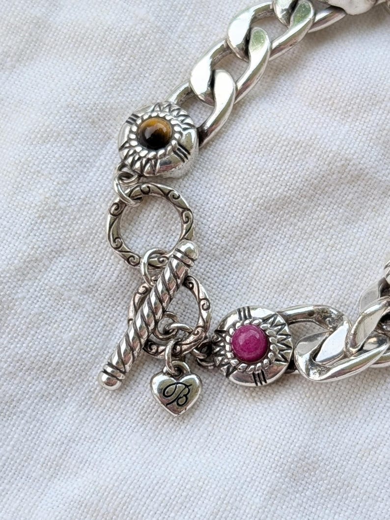 May include: Silver-toned bracelet with a chunky chain design. The bracelet features decorative circular accents with a brown and a pink stone, and a heart charm with the letter "B". The clasp is a toggle style.