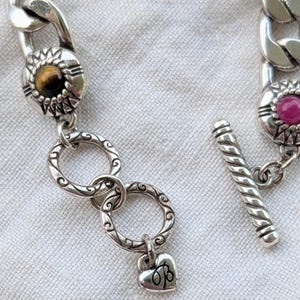 May include: Silver-toned chain bracelet with decorative links. The bracelet features a toggle clasp and decorative links with gemstones. One link has a heart charm with the initials "OB".