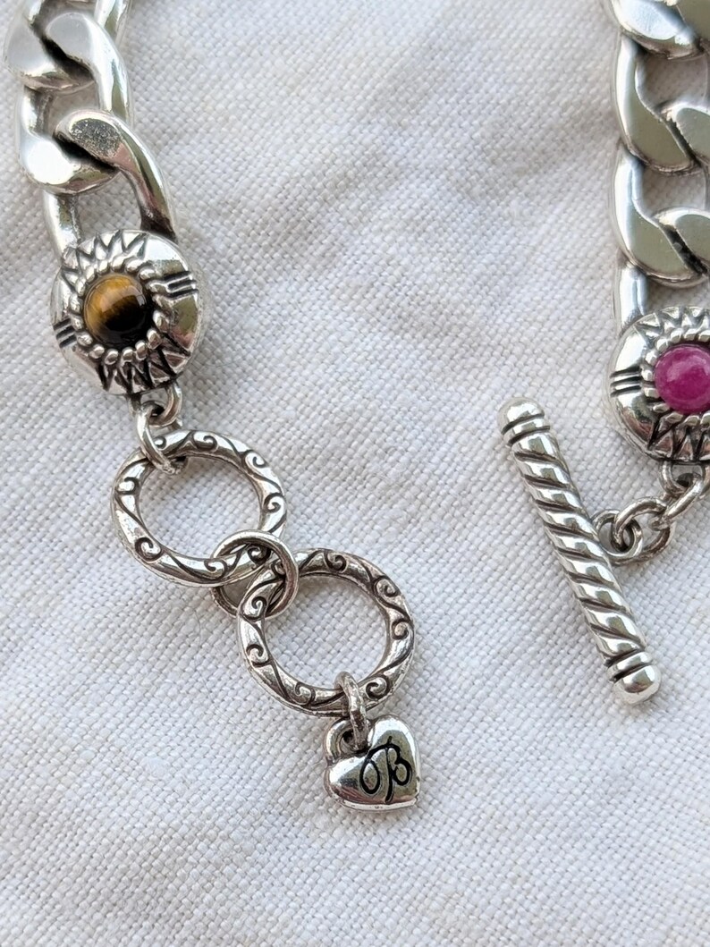 May include: Silver-toned chain bracelet with decorative links. The bracelet features a toggle clasp and decorative links with gemstones. One link has a heart charm with the initials "OB".