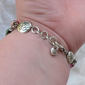 K&ouml;nnte beinhalten: Ein silbernes Kettenarmband mit runden Metallanh&auml;ngern, die mit gravierten Wirbel- und Blumenmustern versehen sind, durchsetzt mit kleinen rosa und gr&uuml;nen Perlen. Ein kleiner herzf&ouml;rmiger Anh&auml;nger mit Gravur h&auml;ngt nahe am Karabinerverschluss.