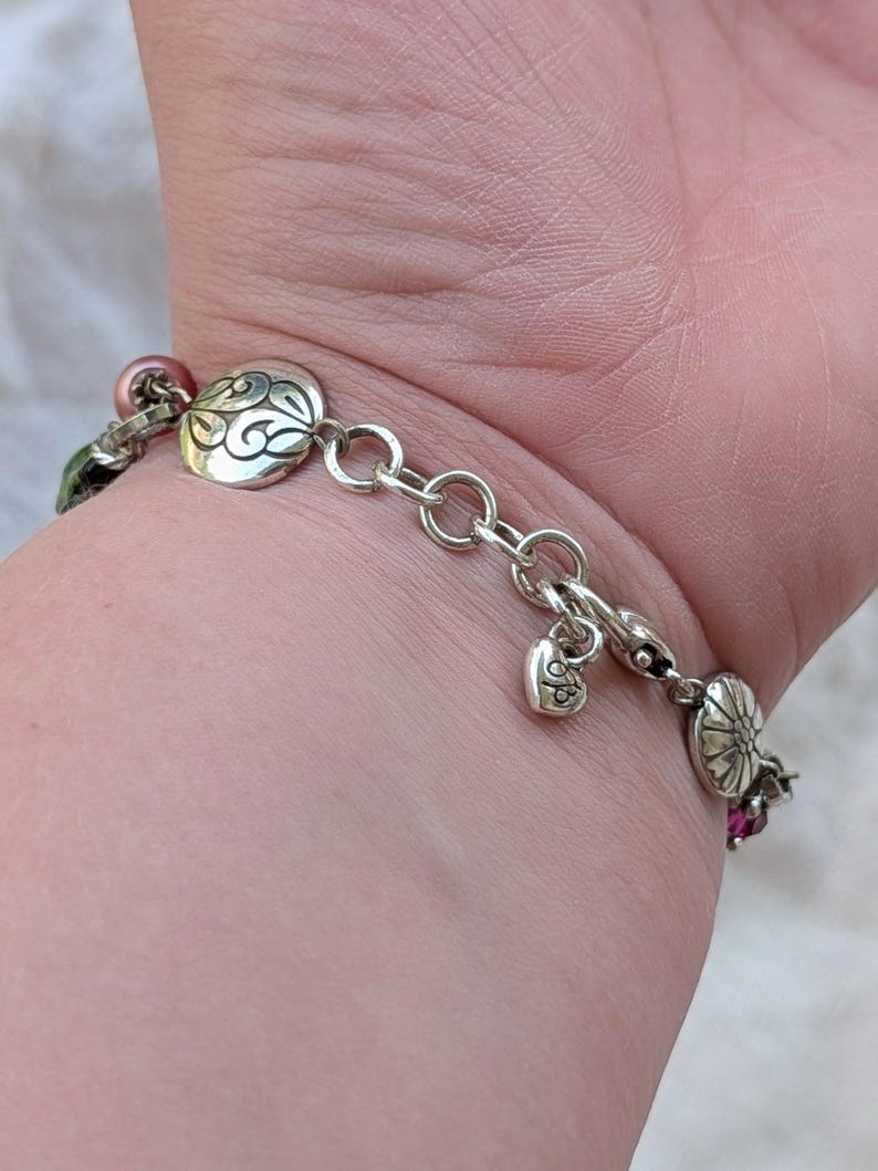 K&ouml;nnte beinhalten: Ein silbernes Kettenarmband mit runden Metallanh&auml;ngern, die mit gravierten Wirbel- und Blumenmustern versehen sind, durchsetzt mit kleinen rosa und gr&uuml;nen Perlen. Ein kleiner herzf&ouml;rmiger Anh&auml;nger mit Gravur h&auml;ngt nahe am Karabinerverschluss.