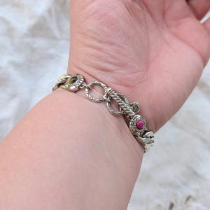 May include: Silver-toned bracelet with intricate floral and rope-like designs. The bracelet features a chain link style with a flower-shaped clasp and a vibrant red gemstone accent. A stylish piece of jewelry.