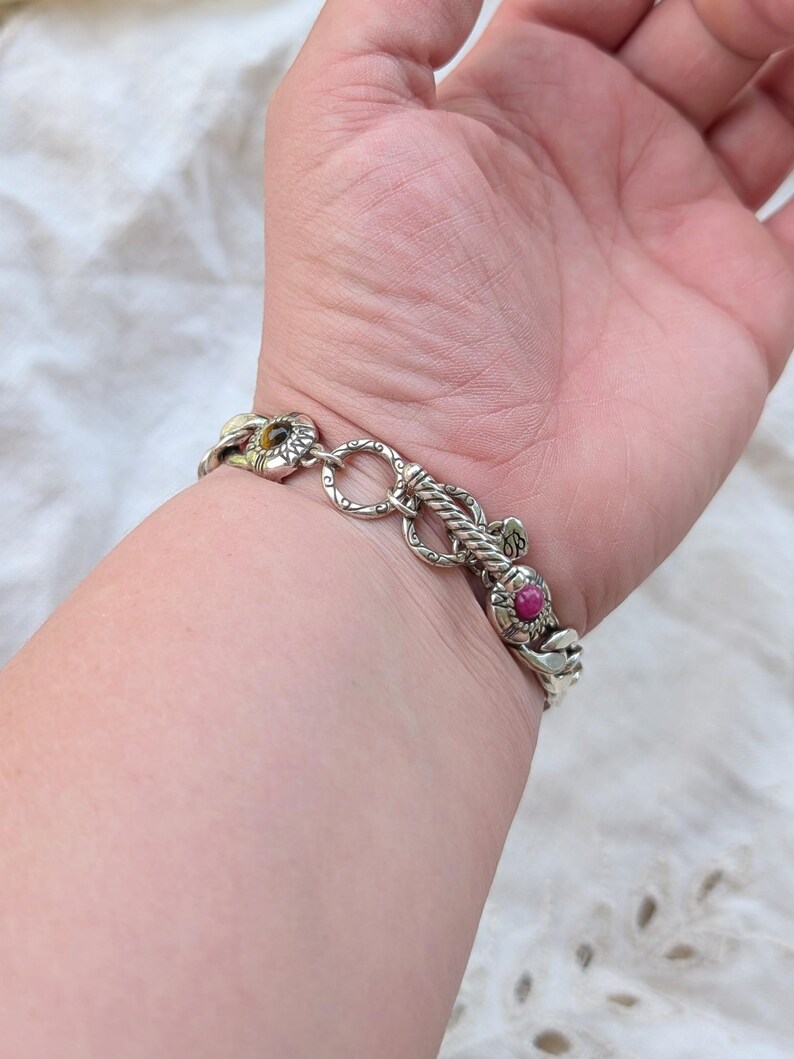 May include: Silver-toned bracelet with intricate floral and rope-like designs. The bracelet features a chain link style with a flower-shaped clasp and a vibrant red gemstone accent. A stylish piece of jewelry.