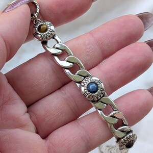 May include: Silver-toned chain bracelet with three decorative circular accents. Each accent features a central gemstone in shades of brown and blue, surrounded by a detailed sunburst design. The bracelet is held against a soft, white background.