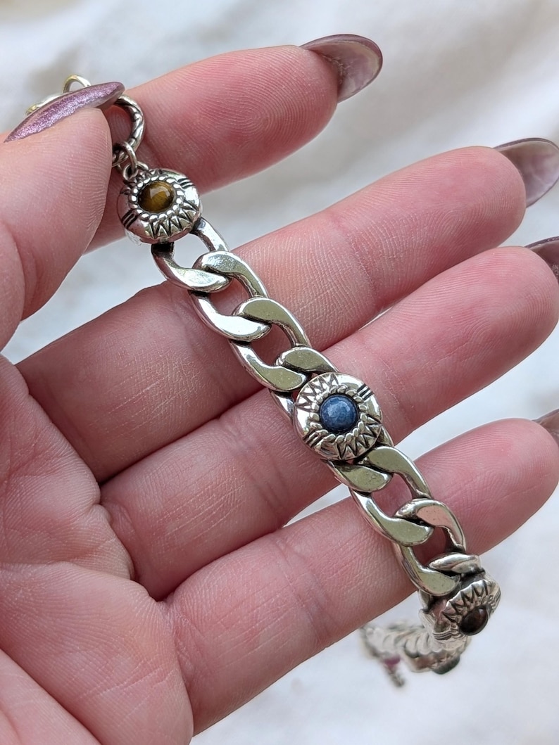 May include: Silver-toned chain bracelet with three decorative circular accents. Each accent features a central gemstone in shades of brown and blue, surrounded by a detailed sunburst design. The bracelet is held against a soft, white background.