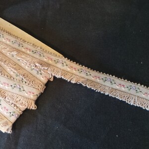 Puede incluir: Un ribete beige con un patr&oacute;n floral y flecos. El ribete est&aacute; sobre una superficie de madera.