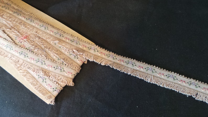 Puede incluir: Un ribete beige con un patr&oacute;n floral y flecos. El ribete est&aacute; sobre una superficie de madera.