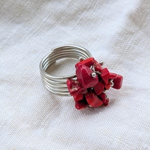 May include: A silver-toned ring with a cluster of bright red coral-like stones. The ring band is made of multiple coiled wires. The coral pieces are clustered together, creating a bold, eye-catching design. The ring is set against a white background.