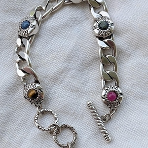 May include: A silver chain bracelet with decorative circular settings, each holding a different colored gemstone. The gemstones include blue, green, red, and brown. The bracelet has a toggle clasp and a heart charm.