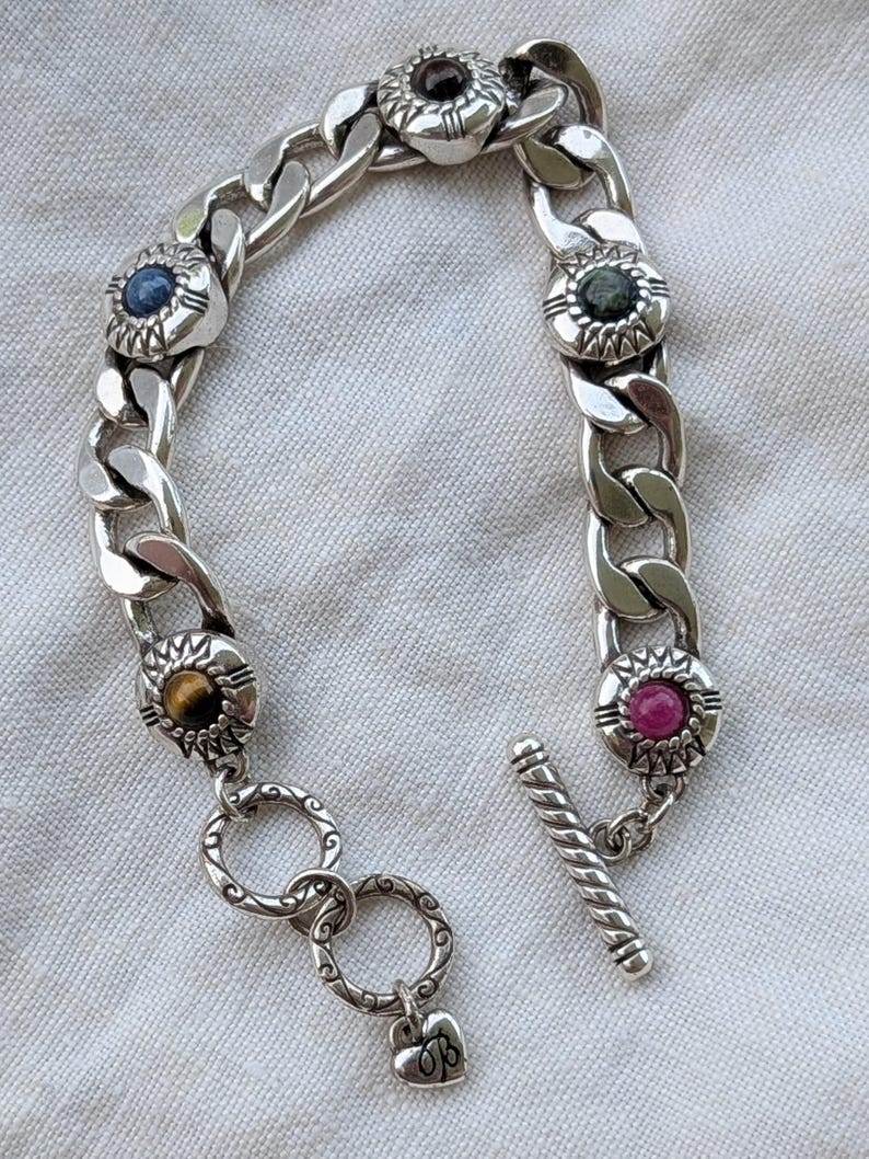 May include: A silver chain bracelet with decorative circular settings, each holding a different colored gemstone. The gemstones include blue, green, red, and brown. The bracelet has a toggle clasp and a heart charm.