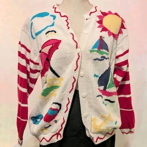 May include: A white long-sleeve knit cardigan featuring a button-front closure and a colorful beach-themed pattern. The design includes a yellow sun, sailboats, a pink umbrella, and red wavy trim. The sleeves have red and white horizontal stripes.