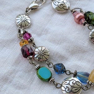 K&ouml;nnte beinhalten: Ein mehrstr&auml;ngiges silberfarbenes Armband mit einer Vielzahl bunter Glasperlen in Rosa, Gelb, Dunkelrot, T&uuml;rkis und Blau, durchsetzt mit runden Metallanh&auml;ngern, die mit Blumen- und Wirbelmustern gepr&auml;gt sind, sowie einem Karabinerverschluss.
