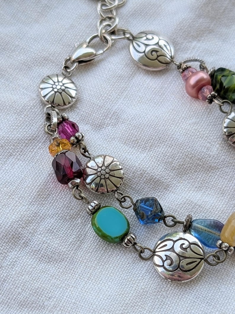 K&ouml;nnte beinhalten: Ein mehrstr&auml;ngiges silberfarbenes Armband mit einer Vielzahl bunter Glasperlen in Rosa, Gelb, Dunkelrot, T&uuml;rkis und Blau, durchsetzt mit runden Metallanh&auml;ngern, die mit Blumen- und Wirbelmustern gepr&auml;gt sind, sowie einem Karabinerverschluss.
