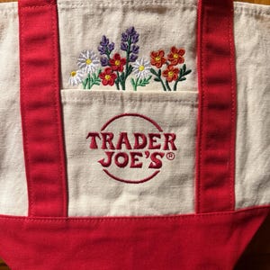 Peut inclure: Un sac fourre-tout en toile couleur crème avec des anses et une base en tissu rouge. L'avant présente un motif floral brodé au-dessus d'une poche avec le texte rouge TRADER JOE'S dans un cercle. Les fleurs sont blanches, rouges et violettes.