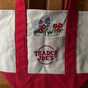 May include: A cream-colored canvas tote bag with red fabric handles and a red base. The front features an embroidered design of colorful wildflowers above a front pocket that displays the red embroidered text Trader Joe's inside a red circle.