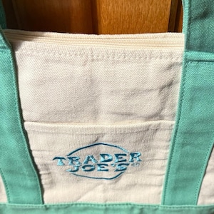 May include: A cream-coloured canvas tote bag featuring mint green handles and a matching base. The bag includes a top zip closure and an exterior front pocket embroidered with the blue text Trader Joe's inside an oval outline design.