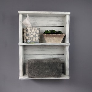 May include: A rustic, white-washed wooden shelf with two levels. The top shelf holds a tissue box with a floral pattern and a small potted succulent. The bottom shelf displays a folded gray towel. The shelf is mounted on a gray wall.