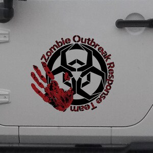 Puede incluir: Un Jeep blanco con una calcomanía negra y roja en la puerta lateral. La calcomanía dice "Zombie Outbreak Response Team" con un símbolo de riesgo biológico y una huella de mano ensangrentada.
