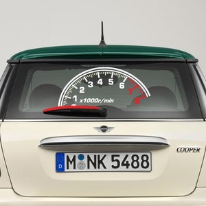 Può includere: Una Mini Cooper bianca con tetto verde. Il lunotto posteriore ha un adesivo di un tachimetro con un ago rosso. La targa è "M NK 5488".