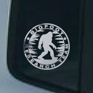 May include: A white vinyl sticker with a black background featuring a silhouette of Bigfoot walking through a forest. The sticker reads "Bigfoot Research Team" in a circle around the silhouette.